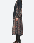 A model is shown from the side wearing a long-sleeved, dark charcoal dress with an intricate multicolor paisley and geometric pattern on the sleeves and skirt, paired with black leather boots against a light gray background.