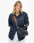 A woman wearing a dark blue denim shirt and white pants carries a dark woven shoulder bag with a brown strap featuring the brand name 'cvc'.