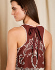 A woman shown from the back wearing a sleeveless burgundy dress with white floral embroidery and a tied bow at the neckline, set against a soft beige curtain background.