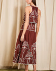 A woman modeling a sleeveless halter-neck midi dress in deep burgundy with intricate white floral embroidery, shown from a rear three-quarter view against a beige curtain backdrop.
