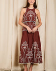 A woman modeling a sleeveless maroon maxi dress with intricate white embroidery, standing against a beige curtain backdrop.