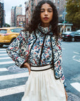 Woman in a floral blouse and white skirt standing on a city street.