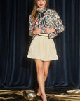 Woman wearing a floral blouse and beige skirt against a dark blue curtain, full body view