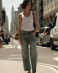 Woman standing on a city street wearing a white tank top and blue jeans.