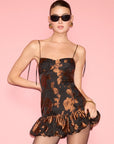 A model wearing a black and brown floral jacquard mini dress with thin straps and a ruffled hem, styled with black sunglasses, gold hoop earrings, and a gold bracelet against a solid pink background.