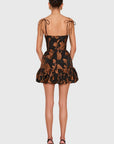 A model viewed from the back wearing a short black dress with a brown floral jacquard pattern, thin shoulder tie straps, and a ruffled hem, paired with black high heels against a light gray background.
