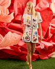 Woman in a colorful embroidered outfit standing in front of a large red floral arrangement.