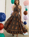 A woman models a halter-neck midi dress with a black base and brown paisley pattern, standing in a studio decorated with colorful paper lanterns.