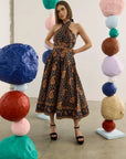 A woman stands in a studio wearing a dark halter-neck midi dress with a brown paisley pattern. The background features colorful sculptural paper ball columns.