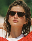 A close-up of a woman with wet hair wearing tortoiseshell sunglasses and a red shirt with a white collar, set against a blurred green and white background.