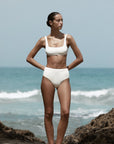 Woman in a white bikini standing on a rocky beach with ocean in the background