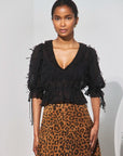 A model wearing a black ruffled short-sleeve blouse with tie details and a brown leopard-print skirt, standing against a light gray background.