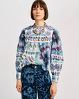 A model wearing a long-sleeve patterned blouse with blue, purple, and white floral motifs, paired with dark blue patterned trousers, against a light gray studio background.