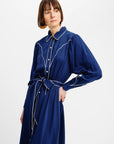 A woman wearing a long-sleeved navy blue shirt dress with white contrast stitching, a pointed collar, and a matching tie belt, photographed against a plain light gray studio background.