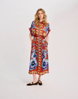 A woman modeling a long-sleeved, midi-length dress with a vibrant red base and a colorful floral and geometric print in blue, purple, yellow, and orange, paired with dark brown flat shoes, against a plain white studio background.