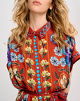 A close-up of a woman wearing a short-sleeve collared dress with a vibrant orange-red base and a multicolor floral print featuring yellow, blue, purple, and green flowers.