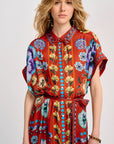 A woman modeling a short-sleeve, tie-neck dress with a vibrant red-orange base and a colorful floral print in blue, purple, yellow, and green, accented with turquoise buttons down the front.