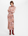 A model wearing a long-sleeved mock-neck midi dress with a colorful geometric and tribal-inspired pattern in pink, orange, red, and black, paired with dark red ankle boots, against a plain white studio background.