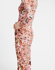 A model shown from the side wearing a long-sleeve, high-neck maxi dress with a dense botanical and paisley print in pink, orange, green, and black on a white background.