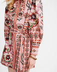 A close-up of a model wearing a pink long-sleeve shirt dress with a bold floral and geometric print in red, green, and black, featuring a tie waist belt.