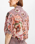 A woman shown from the back wearing a short-sleeve button-front blouse with a mixed floral and gingham print in pink, red, green, and beige tones, paired with a matching gingham skirt against a plain light background.