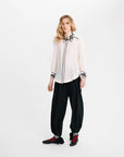 A model stands against a plain white studio background wearing a white long-sleeve blouse with black embroidered trim, black wide-leg pants, and black shoes with red accents.