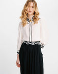 A woman modeling a sheer off-white blouse with black embroidered trim and a black skirt with a black-and-white patterned belt, against a plain white studio background.