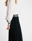 A woman shown from the back wearing a white blouse with black embroidered trim on the cuffs and a black skirt with a black and white patterned waistband, against a plain white studio background.