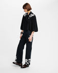 A model wearing a black long-sleeve blouse with white baroque-style embroidery on the shoulders and a matching black wide-leg trouser set with white embroidery on the cuffs, paired with dark brown leather loafers, against a plain white studio background.