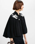 A woman shown in profile wearing a black cropped blouse with dramatic puffed sleeves and white baroque-style embroidery on the shoulders, paired with dark trousers against a plain light background.