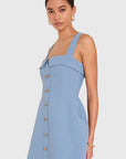 A woman modeling a light blue sleeveless mini dress with a structured, slightly draped bodice, square neckline, and a vertical row of gold buttons down the front, set against a plain light gray background.