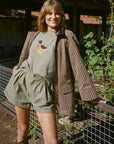 A woman stands outdoors in a garden setting wearing a brown and beige vertically striped blazer, an olive green t-shirt with a duck graphic, matching olive green shorts, and olive green rain boots.
