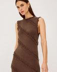 A woman modeling a sleeveless brown knit dress with diagonal fringe details, photographed against a plain light gray studio background.
