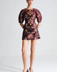 A model wearing a short puff-sleeve dress with a dark burgundy floral pattern and gold buttons, paired with black pointed-toe heels, against a light gray studio background.