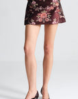 A model's legs wearing a dark brown satin mini skirt with a pink and green floral pattern, paired with dark brown pointed-toe kitten heel pumps, against a light gray studio background.