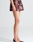 A model's lower body wearing a dark brown floral jacquard mini skirt and black pointed-toe kitten heel pumps, photographed against a clean light gray studio background.