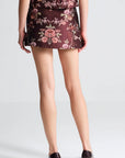 Rear view of a model wearing a short pleated skirt in a dark burgundy brocade fabric with a pink and green floral pattern, paired with dark brown pointed-toe heels.