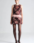 A model stands against a light gray studio background wearing a sleeveless dark burgundy mini dress with a pink floral print, a light pink feathered waistband, sheer black tights, and dark pointed-toe heels.