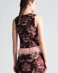 A woman shown from the back wearing a sleeveless dark burgundy mini dress with a pink floral brocade pattern and a light pink feather trim at the waist, set against a plain light gray studio background.