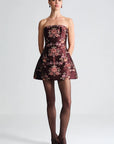A model stands against a light gray studio background wearing a strapless mini dress with a dark burgundy base and an ornate floral jacquard pattern in pink, gold, and beige. She is styled with sheer black tights and dark pointed-toe heels.