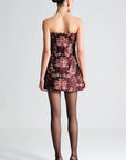 A woman shown from the back wearing a strapless mini dress with a burgundy and pink floral pattern, sheer black tights, and high heels against a light gray studio background.