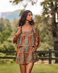 A woman stands outdoors in a grassy park wearing a bohemian-style two-piece outfit with a rust-orange base and blue floral patterns, featuring a matching short dress and a fringed shawl.