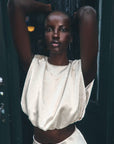 A model with a shaved head poses against a dark green door, wearing a cream-colored, textured two-piece set consisting of a cropped top and matching skirt.