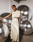 A model wearing a cream satin two-piece set consisting of a cropped sleeveless top and a matching high-waisted skirt, posing in front of large reflective silver mirrors in an art gallery setting.