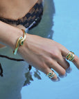 A close-up lifestyle shot of a person's hand and wrist wearing gold snake-themed jewelry, including a wrap-around bracelet and matching rings, set against a blue water background.