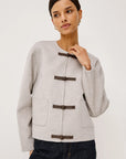model wearing an oat colored jacket with toggle closures and jeans on a light grey background