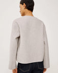 model wearing an oat colored jacket with toggle closures and jeans on a light grey background, back view