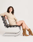 Woman sitting on a modern chair wearing a beige sweater and brown boots against a white background
