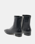 A pair of black leather ankle boots with a side zipper, shown from a rear three-quarter angle against a light gray studio background.