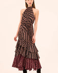 A model wearing a sleeveless halter-neck maxi dress with a black and beige abstract pattern on the bodice and a dark burgundy and beige patterned layered ruffle hem, paired with black ankle boots, against a solid light beige background.